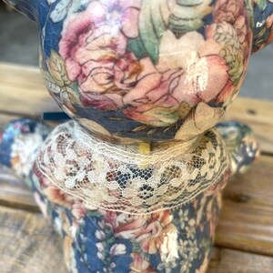 May include: A ceramic teddy bear figurine with a blue and floral patterned design. The bear is wearing a white lace collar.