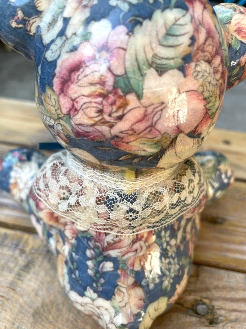 May include: A ceramic teddy bear figurine with a blue and floral patterned design. The bear is wearing a white lace collar.