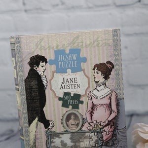 May include: A vintage-style jigsaw puzzle box featuring a couple in period clothing. The box is decorated with a floral pattern and the text "Jigsaw Puzzle Jane Austen 500 Pieces".