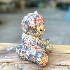 May include: A ceramic teddy bear figurine with a floral pattern in shades of blue, pink, and yellow. The bear is wearing a white lace collar.