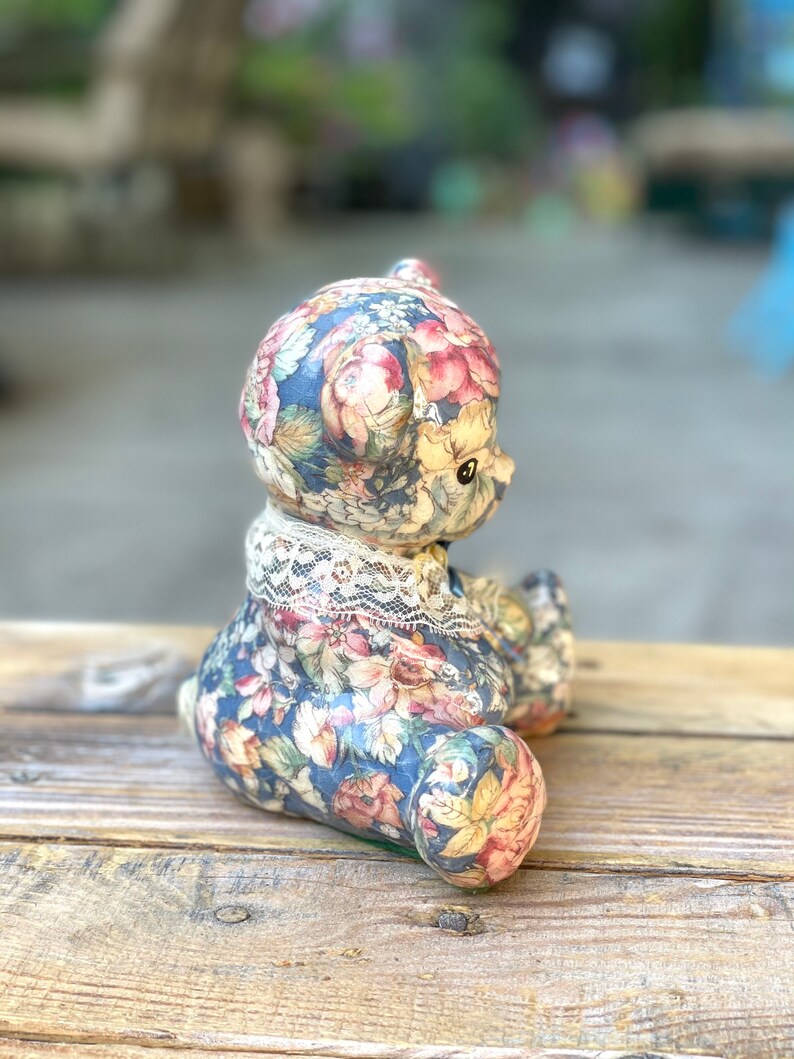 May include: A ceramic teddy bear figurine with a floral pattern in shades of blue, pink, and yellow. The bear is wearing a white lace collar.