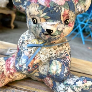 May include: A ceramic teddy bear figurine with a floral pattern in shades of blue, pink, and green. The bear is wearing a white lace collar with a blue ribbon.