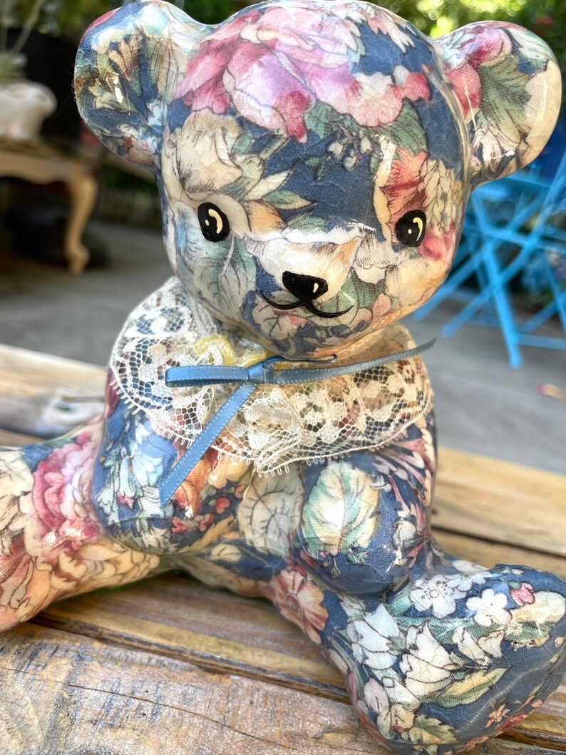May include: A ceramic teddy bear figurine with a floral pattern in shades of blue, pink, and green. The bear is wearing a white lace collar with a blue ribbon.