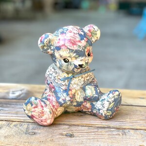 May include: A ceramic teddy bear figurine with a floral pattern in shades of blue, pink, and white. The bear is sitting with its paws crossed and has a blue ribbon around its neck.