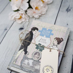 May include: A vintage-style jigsaw puzzle featuring a scene from Jane Austen's novel, with a 500-piece count. The puzzle is packaged in a box with a cream background and a blue and white striped pattern. A brown twine bow is tied around the box, and a cream-colored tag with a gold gear design is attached. The tag reads "So Vintage Good" and includes a website address.