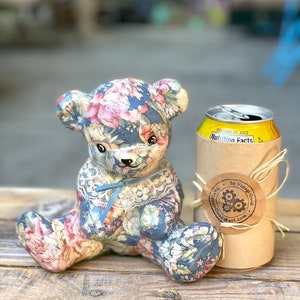 May include: A floral patterned teddy bear sitting next to a can of soda with a brown paper bag wrapped around it. The bag has a sticker that says "So Vintage Good" with a gear design.