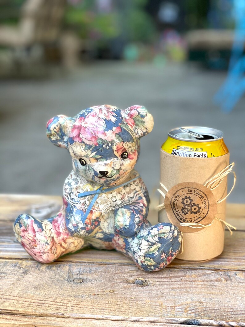 May include: A floral patterned teddy bear sitting next to a can of soda with a brown paper bag wrapped around it. The bag has a sticker that says "So Vintage Good" with a gear design.