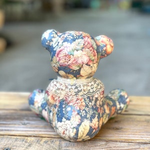 May include: A ceramic teddy bear figurine with a floral pattern in shades of blue, pink, and yellow. The bear is sitting on a wooden surface.