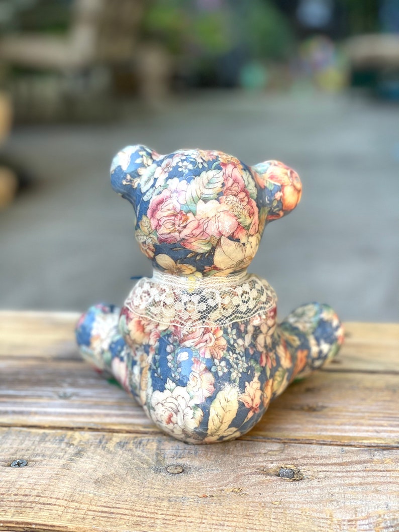 May include: A ceramic teddy bear figurine with a floral pattern in shades of blue, pink, and yellow. The bear is sitting on a wooden surface.