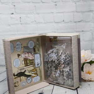 May include: An open box with a vintage-style illustration of a couple on the inside. The box contains a jigsaw puzzle with a plastic bag. The box is on a wooden surface.