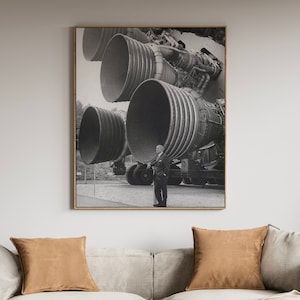 May include: A black and white photograph of a man standing in front of a large rocket engine. The engine is made up of several large, cylindrical sections, each with a series of rings around it. The man is wearing a suit and tie, and he is looking at the camera.