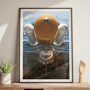 May include: A framed print of a space shuttle, featuring a large orange fuel tank and two white booster rockets. The shuttle is positioned vertically, showcasing its intricate details and design. The print is displayed on a wooden cabinet.