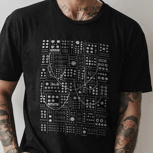 以下が含まれることがあります： モジュラーシンセサイザーパネルの白いグラフィックが特徴の黒いTシャツ。デザインには、さまざまなノブ、ボタン、接続ワイヤーが含まれており、詳細で技術的な美しさを生み出しています。シャツはクラシックなクルーネックと半袖です。
