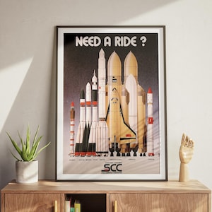 May include: Framed poster featuring various rockets and spacecraft, with the text "NEED A RIDE ?" at the top. The poster has a black frame and is displayed on a wooden cabinet. A small potted plant and a wooden hand sculpture are also on the cabinet.
