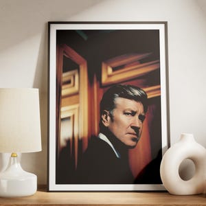 May include: Framed portrait of a man in a dark suit, with a white shirt and tie. The portrait is in a black frame, and is set on a wooden surface. A white lamp and vase are also on the surface.