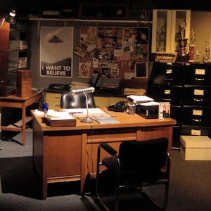 May include: A brown wooden desk with a black leather chair and a lamp. The desk has a telephone, a notepad, and a few papers on it. There is a bulletin board behind the desk with various papers and photos pinned to it. The text on the bulletin board reads "I want to believe".