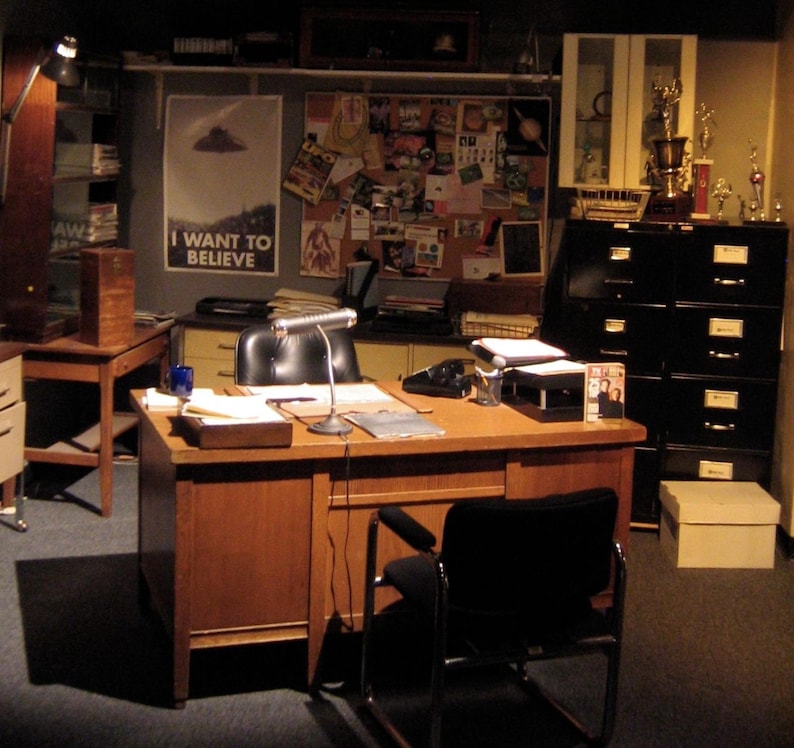 May include: A brown wooden desk with a black leather chair and a lamp. The desk has a telephone, a notepad, and a few papers on it. There is a bulletin board behind the desk with various papers and photos pinned to it. The text on the bulletin board reads "I want to believe".