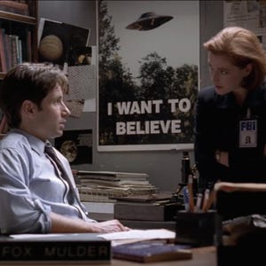 May include: A man and a woman are sitting at a desk. The man is wearing a light blue shirt and a dark tie. The woman is wearing a black suit jacket and an FBI badge. The man has a nameplate that reads "Fox Mulder". There is a poster on the wall behind them that reads "I Want To Believe".