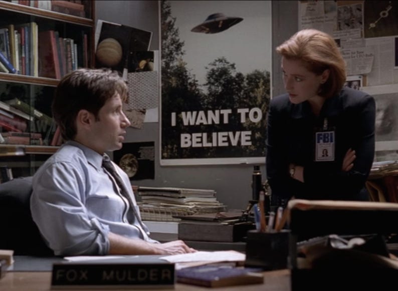 May include: A man and a woman are sitting at a desk. The man is wearing a light blue shirt and a dark tie. The woman is wearing a black suit jacket and an FBI badge. The man has a nameplate that reads "Fox Mulder". There is a poster on the wall behind them that reads "I Want To Believe".