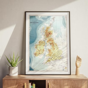 Puede incluir: Mapa antiguo enmarcado de las Islas Británicas, con topografía detallada en tonos amarillos y marrones sobre un fondo azul y blanco. El mapa está en un marco negro, apoyado en un mueble de madera, con una planta y una escultura de mano de madera.