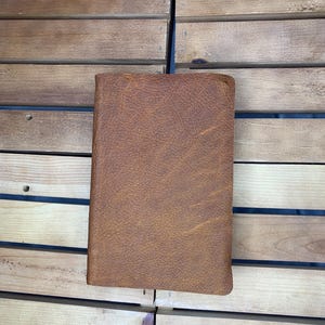 May include: A closed, rectangular, brown leather journal with a textured surface. The journal is positioned vertically against a wooden background. The leather has a natural, aged appearance.