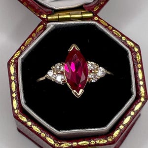 May include: A gold ring featuring a central marquise-cut deep red gemstone flanked by three small clear round stones on each side, presented inside an octagonal dark red jewelry box with gold trim and a white interior lining.