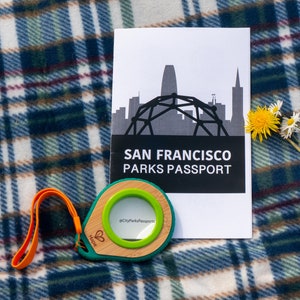 May include: A green and brown park passport holder with a clear magnifying lens and a black and white pamphlet that says "San Francisco Parks Passport" with a graphic of the city skyline and a dome structure.
