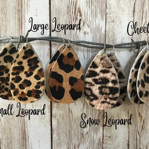 May include: Five pairs of teardrop-shaped leather earrings with different animal print patterns. The earrings are labeled "Large Leopard", "Small Leopard", "Snow Leopard", "Cheetah", and "Leopard".