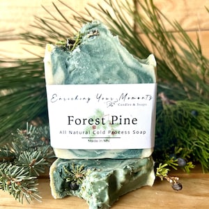 May include: A bar of green and white soap with a label that reads "Enriching Your Moments Candles & Soaps Forest Pine All Natural Cold Process Soap Made in MN".
