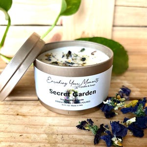 May include: A round, gold-colored candle tin with a lid, labeled "Secret Garden." The candle is filled with white wax and decorated with dried flowers. The label reads "Enriching Your Moments" and "Soy Wax Blend Candle."