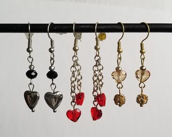 May include: Five pairs of dangle earrings with different styles. Two pairs feature a black bead and a silver heart charm. Two pairs feature a chain with red heart charms. One pair features a gold chain with a brown heart charm and a gold bead.