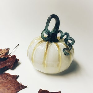 May include: White glass pumpkin with gold accents and a green glass stem. The pumpkin is sitting on a white surface with brown leaves scattered around it.