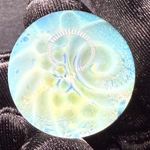 May include: A circular glass art piece with a swirling design in blue, yellow, and white. A silver-coloured, circular pattern is embedded in the centre. The piece is held by a black glove.