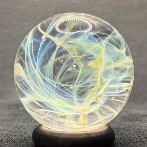 May include: A clear glass sphere with swirling blue and yellow patterns, set on a dark wooden base. The orb is lit from within, producing a glowing effect. The design is artistic and decorative, with a focus on light and colour.