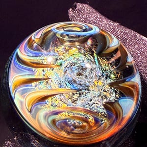 May include: A clear glass marble with swirling patterns in shades of blue, orange, and yellow. The center of the marble features a textured, iridescent design. The marble is set against a dark background.