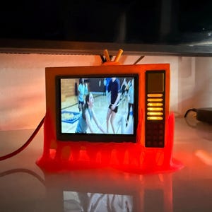 Puede incluir: Un pequeño televisor naranja de estilo retro con una pantalla que muestra un vídeo de personas practicando deportes. El televisor tiene una base roja y una pequeña antena. La pantalla es rectangular y el televisor tiene un panel de control lateral.
