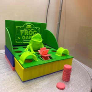 May include: A green plastic tabletop game titled FROG GAME with a central frog figure, a pink waterwheel mechanism, and multiple circular holes. The base is yellow with blue accents, and a stack of small pink circular tokens sits nearby.