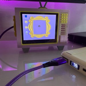 Puede incluir: Un pequeño televisor de estilo retro con una pantalla que muestra una interfaz de escritorio y el icónico marco de Friends. El televisor es blanco y amarillo, con un cable morado conectado a un dispositivo blanco. La pantalla muestra un fondo morado con acentos amarillos.