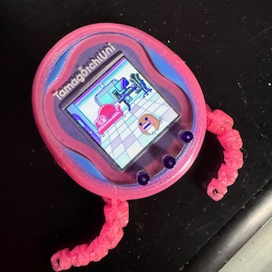 Puede incluir: Un Tamagotchi Uni rosa con un diseño de espiral azul y morado. La pantalla muestra una imagen pixelada de una habitación con muebles y un personaje. El Tamagotchi tiene brazos rosados y rizados impresos en 3D.