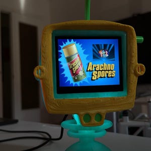 Puede incluir: Un televisor caprichoso de estilo retro, con una base turquesa y un marco de pantalla amarillo y turquesa. La pantalla muestra las palabras "Arachno Spores" y una imagen de un aerosol. Una antena verde se extiende desde la parte superior.