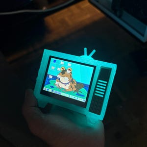 May include: A small, glowing teal-colored television with a screen displaying a cartoon frog. The TV has a retro design with an antenna and a series of vertical lines on the side. The frog is brown and white, wearing a collar and sitting on a lily pad.