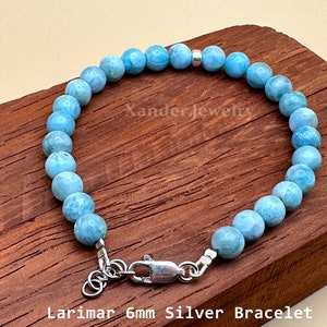 May include: A bracelet featuring round, light blue Larimar beads, each approximately 6mm in diameter, accented with a single silver bead. The piece includes a silver lobster claw clasp and an adjustable chain. Text reads Xander Jewelry.