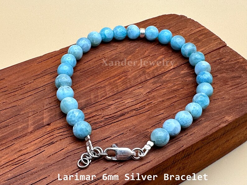 May include: A bracelet featuring round, light blue Larimar beads, each approximately 6mm in diameter, accented with a single silver bead. The piece includes a silver lobster claw clasp and an adjustable chain. Text reads Xander Jewelry.