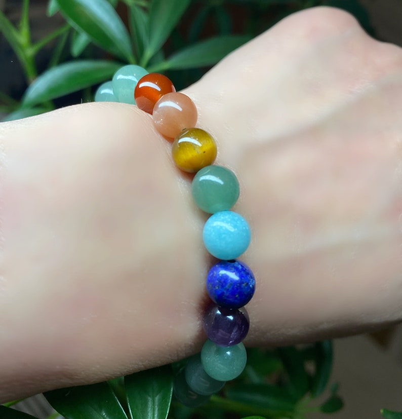 May include: A beaded bracelet featuring a sequence of smooth, polished spherical stones in various colors including red, orange, yellow, green, light blue, dark blue, and purple, arranged in a single strand on an elastic cord.
