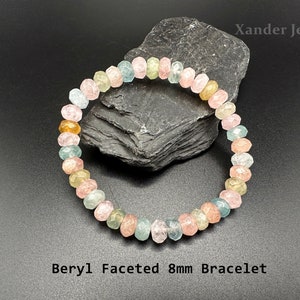 May include: A bracelet made of faceted beryl beads in shades of pink, green, and yellow. The beads are 8mm in diameter and are strung on a clear elastic cord. The bracelet is displayed on a dark grey stone background. The text "Beryl Faceted 8mm Bracelet" is printed below the bracelet.