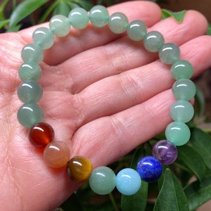 May include: A beaded bracelet featuring a series of smooth, round, light green aventurine beads interspersed with a single dark red, orange, brown, light blue, dark blue, and purple bead, all strung together on a flexible, elastic cord.