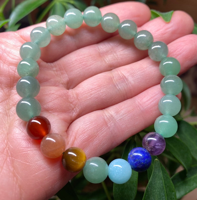 May include: A beaded bracelet featuring a series of smooth, round, light green aventurine beads interspersed with a single dark red, orange, brown, light blue, dark blue, and purple bead, all strung together on a flexible, elastic cord.