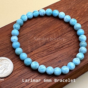 May include: A circular bracelet composed of light blue, marbled Larimar gemstone beads. The beads measure 6mm in diameter. A US dime coin is placed next to the bracelet for scale. The text Xander Jewelry and Larimar 6mm Bracelet is visible.