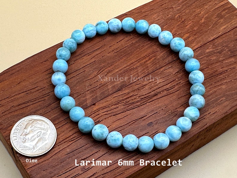 May include: A circular bracelet composed of light blue, marbled Larimar gemstone beads. The beads measure 6mm in diameter. A US dime coin is placed next to the bracelet for scale. The text Xander Jewelry and Larimar 6mm Bracelet is visible.
