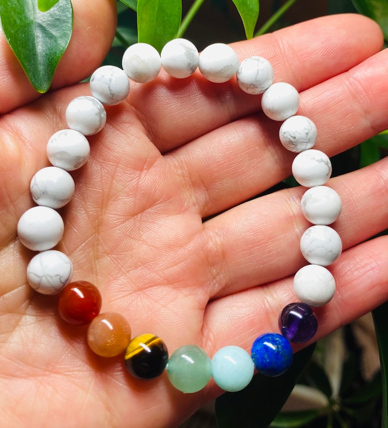 May include: A stretch bracelet composed of round white howlite beads with grey veining, accented by a sequence of seven smooth, polished gemstone beads in red, orange, yellow, green, light blue, dark blue, and deep purple colors.